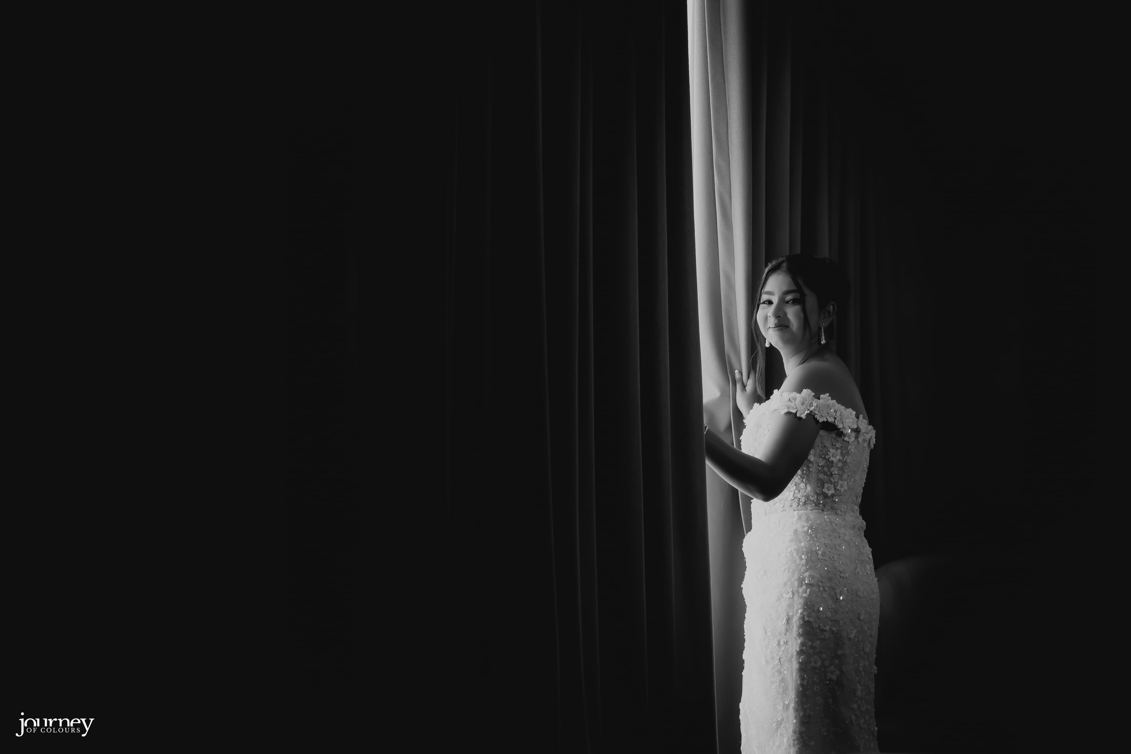 Black and white portrait of a bride opening the curtain as light enters the room